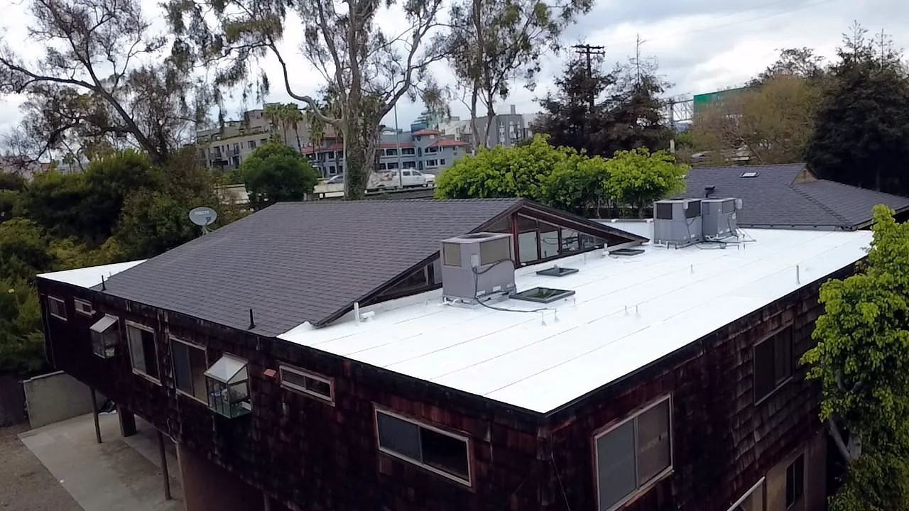 White low-slope membrane, skylights and HVAC units beside a new dark shingle roof section, West Los Angeles